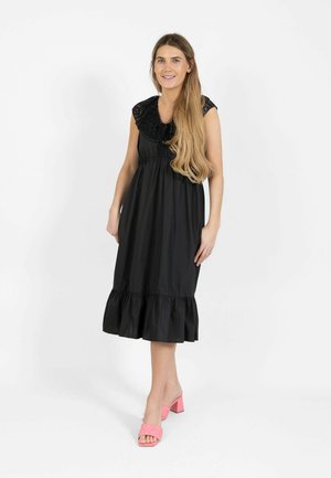 Femme aux longs cheveux portant une robe midi noire sans manches avec un ourlet à volants et des sandales à talons carrés ouverts roses, debout sur un fond blanc.