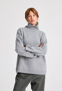 Szürke kötött pulóver magas bordás gallérral, ejtett vállakkal és széles ujjakkal, texturált felülettel és laza fitettel.