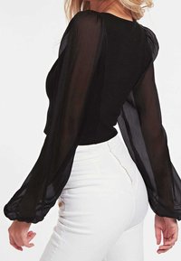 Sort bluse med gennemsigtige, puffede lange ærmer og en tætsiddende krop. Bagsiden er glat og er parret med hvide jeans med høj talje, der har gyldent hardware.