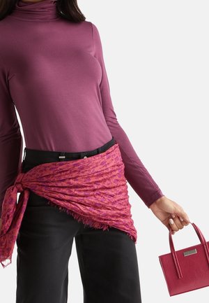 Kobieta w długim rękawie mauve golfa, czarnych spodniach, różowej szaliku w wzory przewiązanym w talii, trzymająca małą czerwoną fakturowaną torebkę za uchwyt.