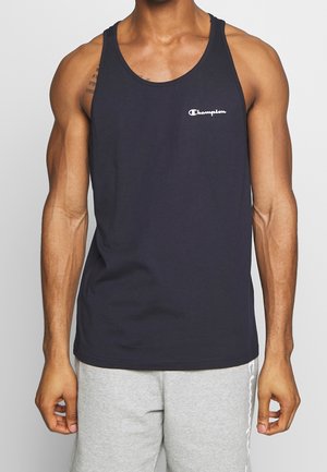 Mann trägt ein schwarzes Champion Tanktop und hellgraue Shorts und steht vor einem einfarbigen hellen Hintergrund.