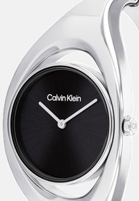 Montre en métal argenté avec un cadran rond noir, affichant un design minimaliste et le logo Calvin Klein. Deux aiguilles blanches indiquent l'heure.