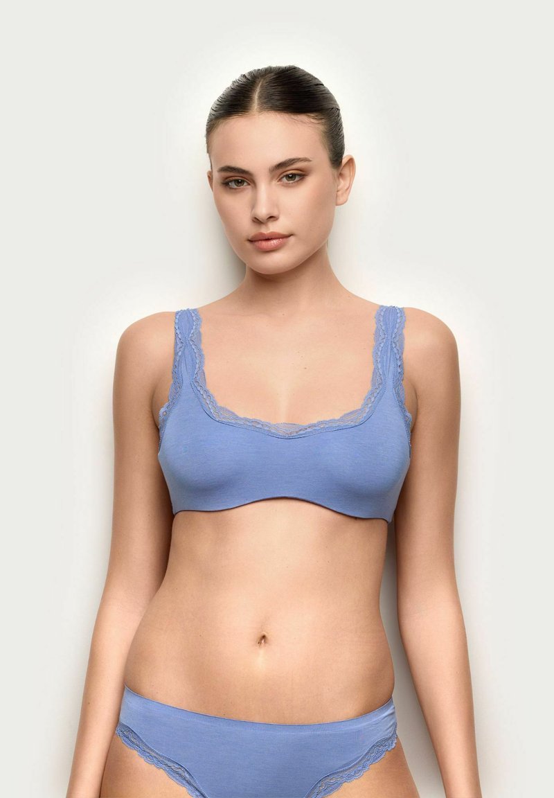 Mujer de cabello oscuro que lleva un conjunto a juego de bralette y bragas azul claro con encaje, de pie contra un fondo claro y liso.