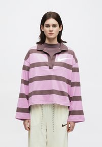 Νέος με ίσια σκούρα μαλλιά, φορώντας ένα βαρετό μπλε και καφέ ριγέ τοπ και ανοιχτούς παντελόνες με λογότυπο Nike.