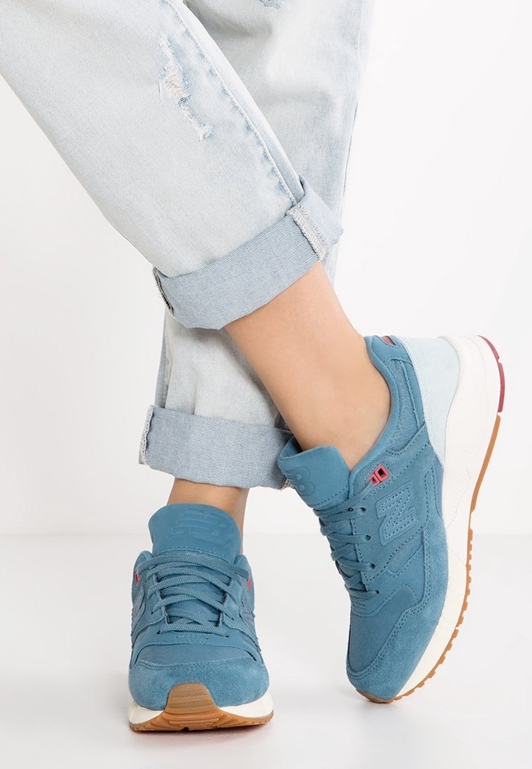 Baskets en daim bleu avec semelles blanches et gomme portées avec un jean bleu clair retroussé, montrant des chevilles croisées sur un fond blanc.