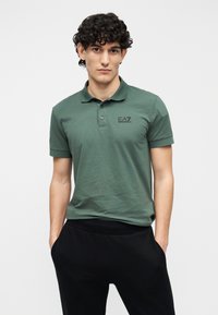 Polo de algodão verde com gola, mangas curtas, pala de dois botões e logotipo EA7 em estampa escura no lado esquerdo do peito. Usado com calças pretas.