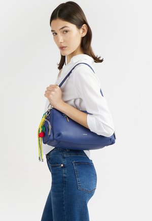 Giovane donna che indossa una camicia bianca e jeans blu, che tiene una borsa in pelle blu con nappe colorate sulla spalla.