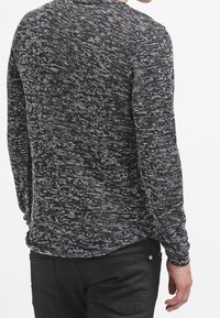 Muž v černobílém melírovaném svetr s dlouhým rukávem a černých kalhotách, zobrazený zezadu s jednou uvolněnou rukou.