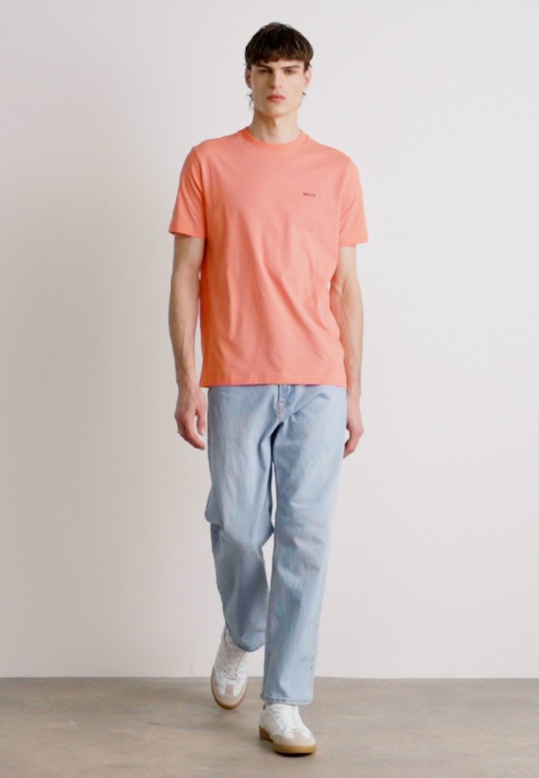 T-shirt à manches courtes de couleur pêche avec un col rond, associé à un jean décontracté bleu clair et des baskets blanches, se tenant sur un sol en béton.
