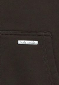 Tissu marron à surface texturée avec une étiquette rectangulaire argentée embossée du texte « bazix republic » sur la poche.