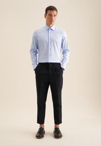 Himmelblaues langärmliges Button-Up-Hemd, kombiniert mit maßgeschneiderten schwarzen Hosen und schwarzen Schuhen, vor einem neutralen Hintergrund stehend.
