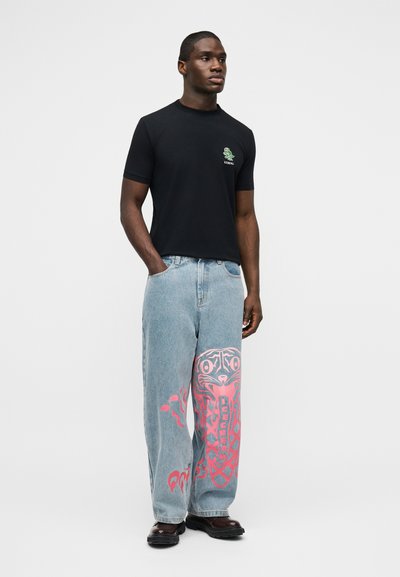Άνδρας με μαύρο κοντομάνικο πουκάμισο με μικρό λογότυπο και ανοιχτό μπλε τζιν παντελόνι με μεγάλο ροζ γραφικό σχέδιο στο αριστερό πόδι, στέκεται μπροστά σε απλό φόντο.