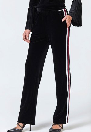 Kvinde iført sorte fløjlsbukser med hvide og røde sidelommer, kombineret med sorte hæle og en sort top med plisserede ærmer.