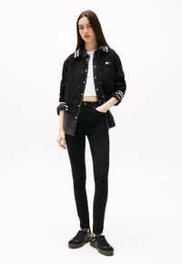 Νεαρή γυναίκα στέκεται με το ένα χέρι στην τσέπη, φορώντας μαύρο varsity μπουφάν, λευκό crop top, μαύρο skinny τζιν και μαύρα αθλητικά παπούτσια.