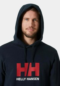 Felpa con cappuccio blu navy, dotata di una tasca a marsupio frontale e un grande logo Helly Hansen rosso e bianco. Tessuto morbido e coulisse regolabile.