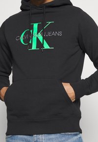 Felpa nera con tasca a marsupio frontale, caratterizzata da un grande logo verde "CK" e "CALVIN KLEIN JEANS" in bianco, con cappuccio regolabile.