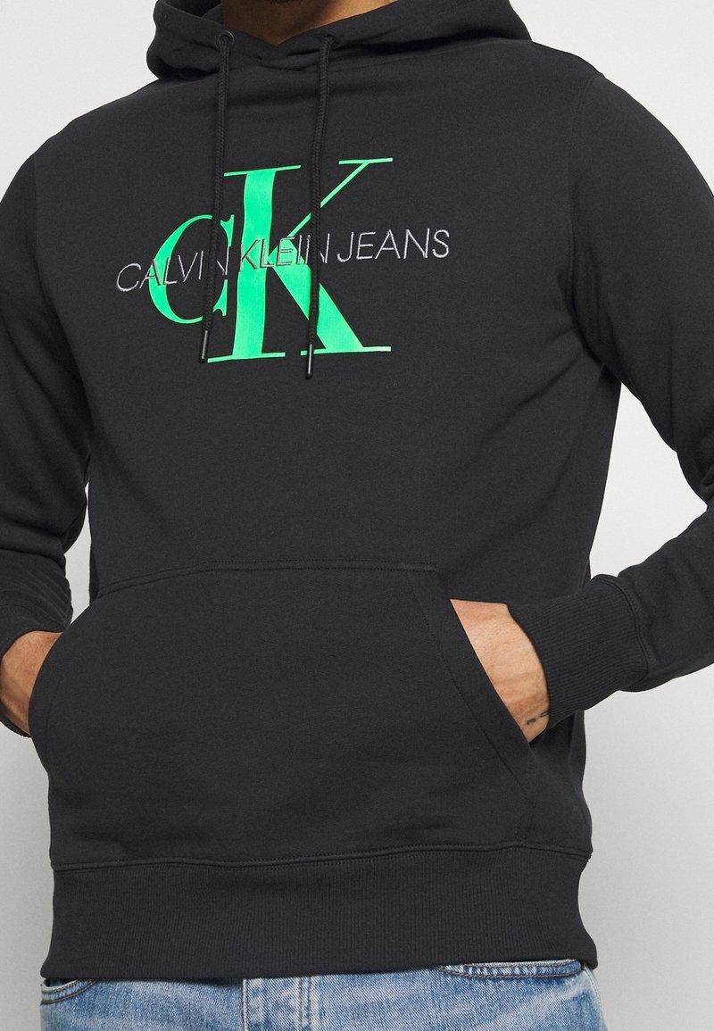 Felpa nera con tasca a marsupio frontale, caratterizzata da un grande logo verde "CK" e "CALVIN KLEIN JEANS" in bianco, con cappuccio regolabile.