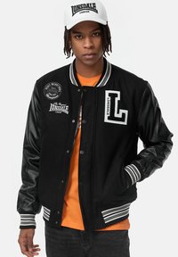 Svart varsity-jacka med läderärmar, vita randiga muddar och krage, LONSDALE-patch på bröstet, buren över en orange tröja.