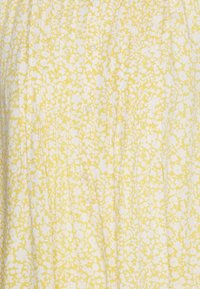 Keltainen kangas, jossa on valkoinen kukkakuosi ja pystyrypytyksiä, keskellä on rivi pieniä beigejä nappeja.