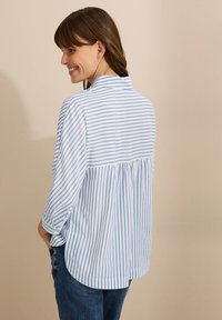 Camisa de rayas azul y blanca con dobladillo curvado, cuello abotonado y detalles fruncidos en la espalda. Las mangas enrolladas muestran un ajuste relajado en una tela suave.