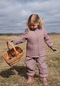 Giacca trapuntata viola e pantaloni abbinati, guanti coordinati e stivali di gomma. Tiene in mano un cesto intrecciato con mele rosse e pigne. Sfondo di campagna.