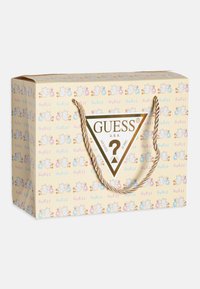 Hnědá papírová dárková taška se zlatými kroucenými uchy. Na tašce se opakují pastelové vzory ptáků a logo "GUESS" ve tvaru trojúhelníku uprostřed.