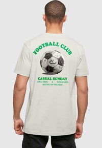 Kurzärmliges Creme-T-Shirt mit einem großen Schwarz-Weiß-Fußball-Graphic auf dem Rücken, mit grünen Schriftzug "Football Club Casual Sunday."