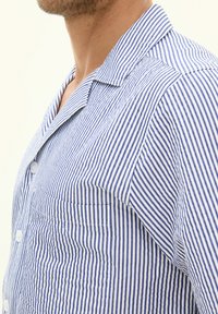 Camicia a righe con linee verticali blu e bianche, chiusura con bottoni e una tasca sul petto. Tessuto morbido con colletto e maniche corte.