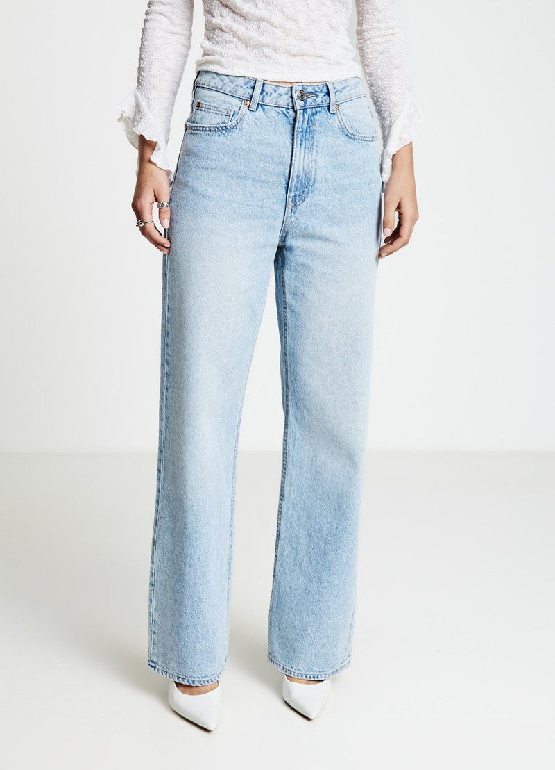 Jean bleu clair à jambes larges, en denim, taille haute, design à cinq poches, assorti à des talons blancs pointus. Légère usure visible.