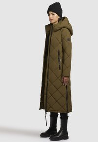 Oliven grøn, lang quilted pufferjakke med hætte, der har sidelommer og et logo-patch. Sammen med sorte ankelstøvler og en strikket hue.