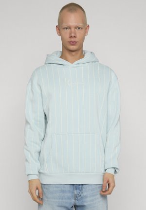 Junge Person trägt einen hellblauen Kapuzenpullover mit dünnen weißen Vertikalstreifen und hellblaue Jeans, steht vor einem grauen Hintergrund.