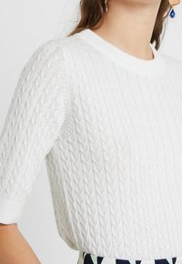 Valkoinen neulepaita, jossa on teksturoitu palmikkokuvio, lyhyet hihat ja pyöreä pääntie. Yksityiskohtina ovat ribbineulottu helma ja hihansuut.