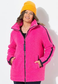 Vaahtomuovinen vaaleanpunainen fleece-takki, jossa on vetskari, korkea kaulus ja sivutaskut; takissa on tummansinisiä raidallisia yksityiskohtia hihoissa ja hihansuissa.