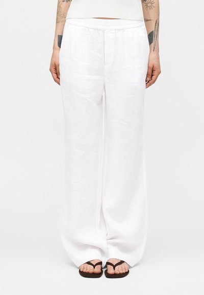 Personne portant un pantalon blanc ample, un débardeur blanc côtelé, des tongs noires, montrant des tatouages sur les avant-bras sur un fond uni.