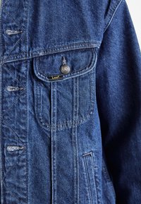 Donkerblauwe spijkerjas met een klassieke snit. Voorzien van een grote voorzak, metalen knoopsluiting en gestikte details. Etiket zichtbaar.