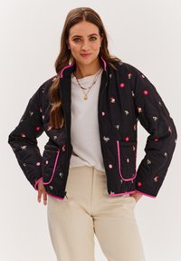 Čierna prešívaná bunda s pestrou kvetinovou výšivkou, ružovým lemovaním a vreckami. Zips a dlhé rukávy. Nosená cez bielu košeľu.