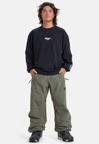 Chemise noire à manches longues avec un logo sur la poitrine, associée à un pantalon cargo olive ample. Des bottes noires ajoutent du contraste et de la praticité.