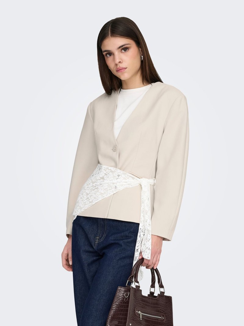 Blazer beige à silhouette ajustée, ceinture en dentelle, manches larges et sac à main marron à motif crocodile. Jean bleu foncé complète la tenue.