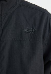 Svart zip-up jacka i texturerat tyg, med en broderad logo i det övre hörnet och en ståkrage.