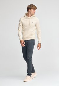 Felpa beige con tasca a marsupio, abbinata a pantaloni grigio scuro e sneakers chiare. Presenta un piccolo logo sul petto.