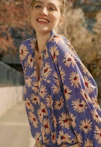Blumenbluse mit blauem Hintergrund, großen cremefarbenen Gänseblümchen und roten Akzenten. Leichter Stoff mit lockerer Passform und Knopfdetails.