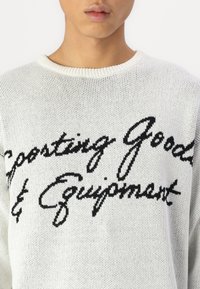 White knit sweater with black cursive text "Sporting Goods & Equipment" across the chest. Textured fabric, round neckline, loose fit.