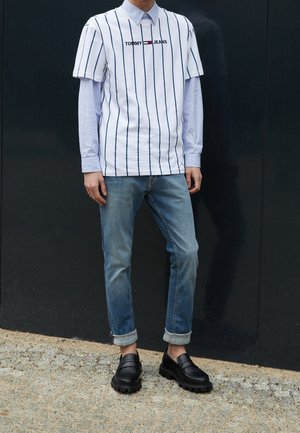 Osoba oblečená v bílé pruhované tričko Tommy Jeans přes světle modrou košili s dlouhým rukávem, ohrnuté modré džíny a černé robustní mokasíny stojící na chodníku.