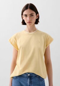 Gelbes T-Shirt mit kurzen Ärmeln, aus weichem Baumwollmaterial, mit einem lockeren Schnitt und einem kleinen, auf der Brust eingestickten Logo. Kombiniert mit Jeans.