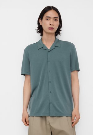 Jeune personne aux cheveux lisses, mi-longs et foncés, portant une chemise boutonnée à manches courtes vert pâle et un pantalon beige, debout contre un mur blanc.