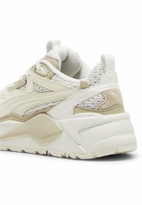 Zapatilla beige gruesa con acentos de malla y ante. Presenta un cuello acolchado texturizado y una suela gruesa y estriada. Diseño elegante y moderno.