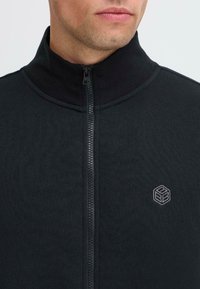 Giacca in fleece nera con zip e colletto alto, caratterizzata da un logo sottile grigio sul lato sinistro del petto e un tessuto testurizzato.
