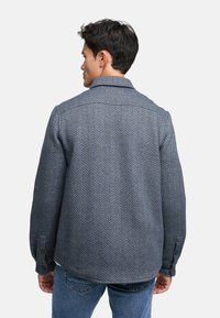 Giacca camicia a motivo spina di pesce in grigio scuro con colletto, maniche lunghe e polsini con bottoni. Il materiale è strutturato e denso.