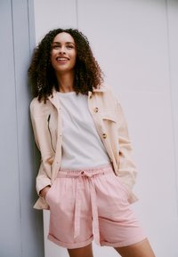 Lys beige jakke med knapper over en hvit ribbestrikket skjorte, kombinert med rosa shorts med snøring, har en avslappet passform og rullede ermer.