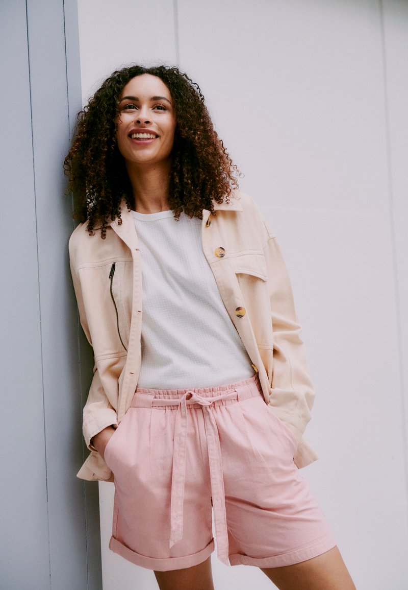 Lys beige jakke med knapper over en hvit ribbestrikket skjorte, kombinert med rosa shorts med snøring, har en avslappet passform og rullede ermer.